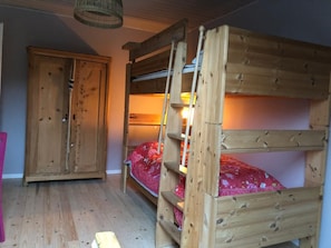 A bunk bed with a ladder, a wooden wardrobe, and a hanging light fixture in a room with wooden flooring.