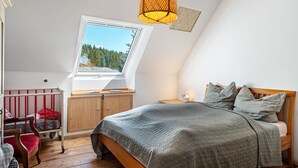 A bedroom with a sloped ceiling, a window with a view of trees, a bed with pillows, a nightstand, and a wooden chair. - Apartment 'Rotkäppchen' with Shared Garden and Wi-Fi (Eisenbach (Hochschwarzwald))