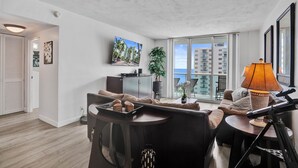 A modern living room with a sofa, a coffee table, a TV, and a balcony with a view of buildings. - Beachfront Getaway | Direct Ocean Views + Balcony (Hollywood)