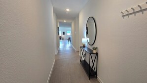 A hallway with a wooden floor, a black console table with a mirror and a plant, and a wall-mounted shelf with hooks. - Cute Brand New Home in McKinney (McKinney)