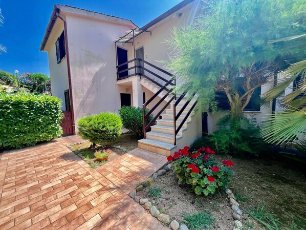 A two-story house with a brick pathway leading to a wooden door, surrounded by lush greenery and a small garden with red flowers.