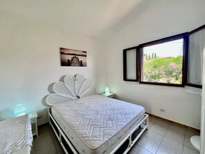 A bedroom with a bed, a nightstand, a lamp, a window with a view of greenery, and a painting on the wall.