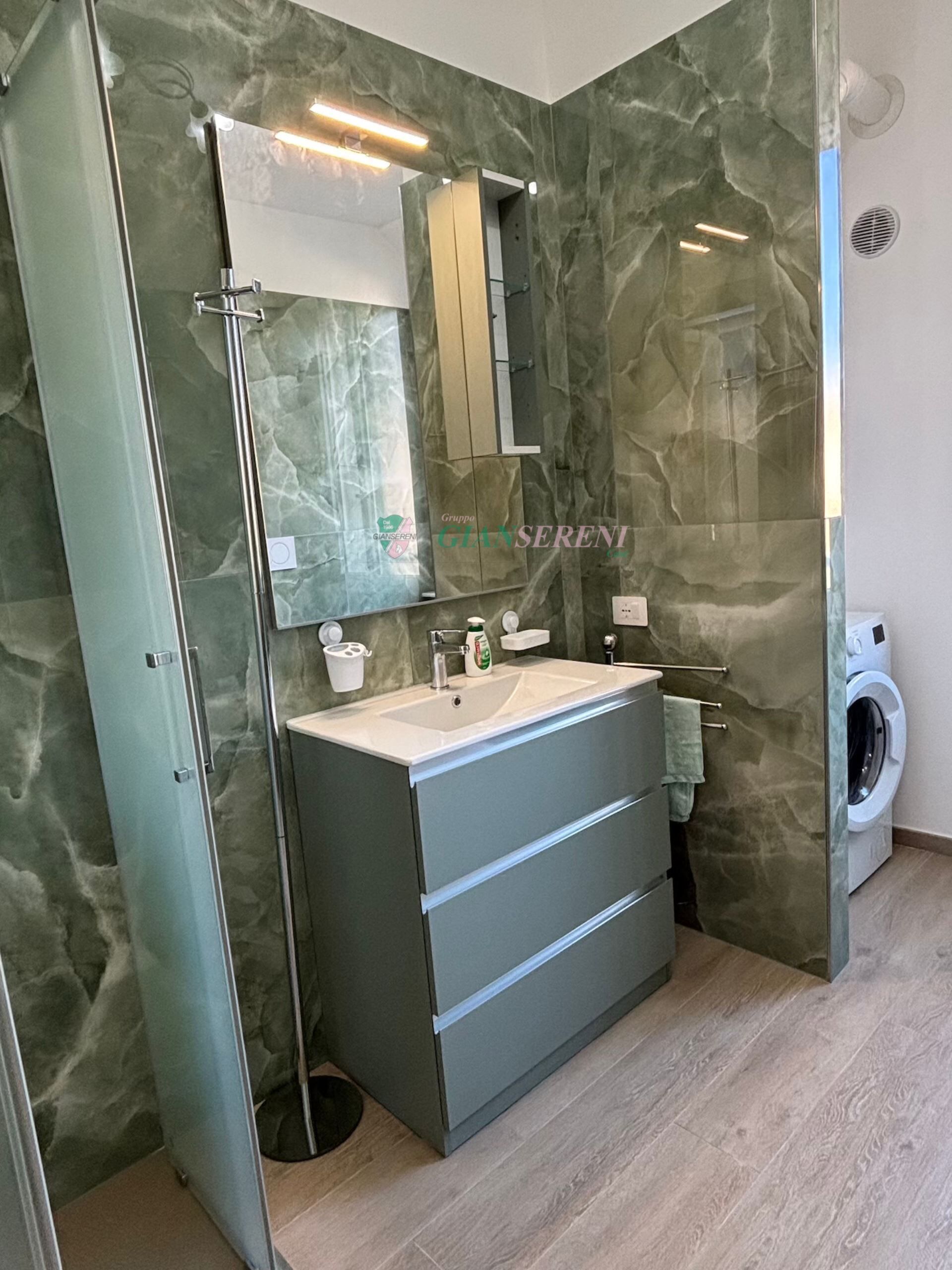 A modern bathroom with a marble backsplash, a glass shower enclosure, a built-in sink, and a washing machine.