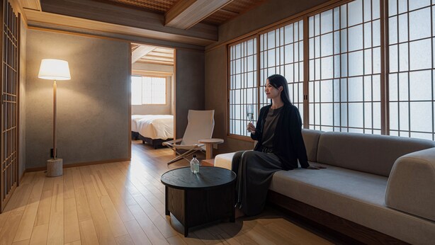 A room with a sofa, a chair, a lamp, a small table, and a bed in the background.