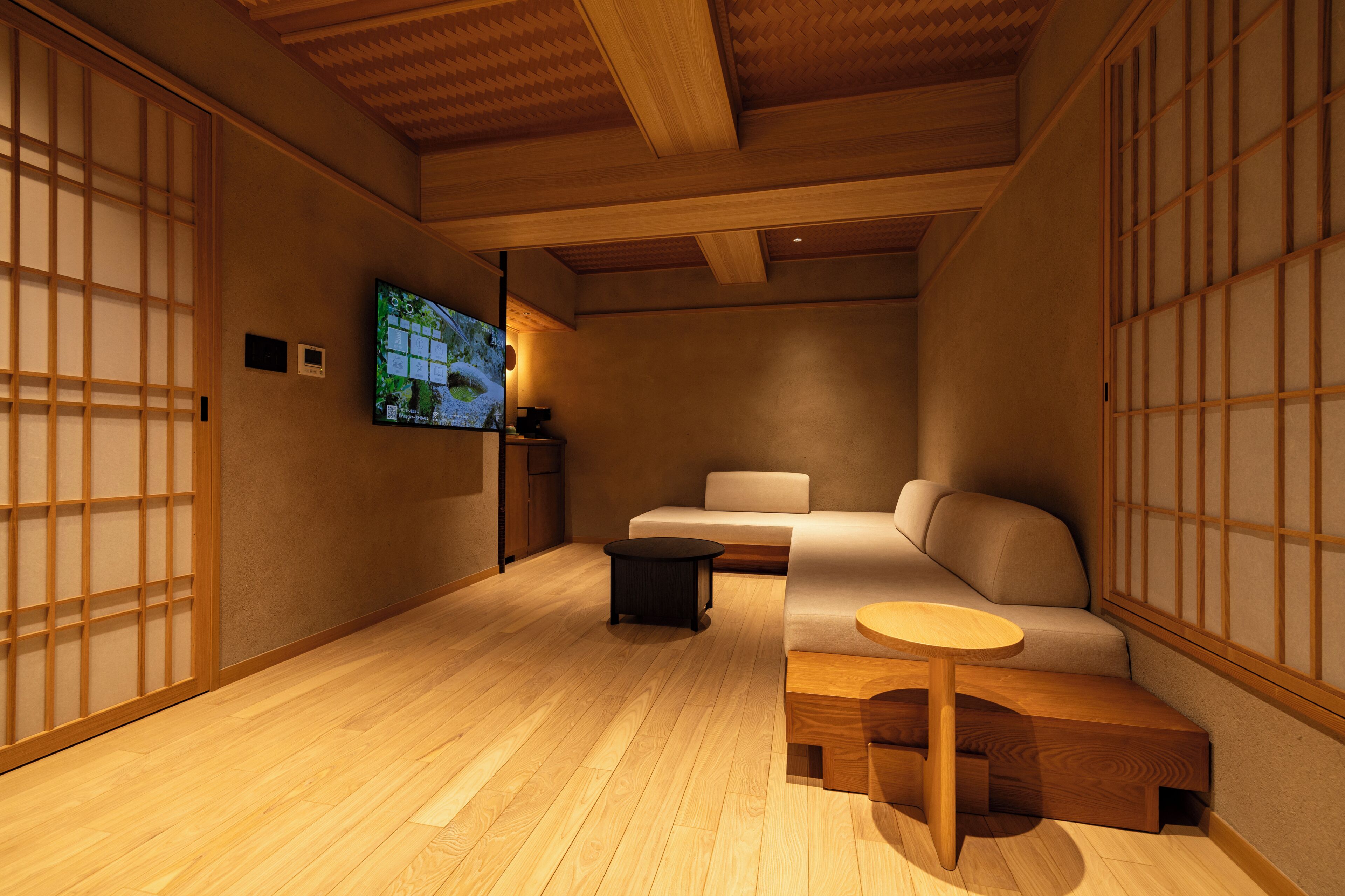 A traditional Japanese-style room with tatami flooring, a low wooden ceiling, sliding doors, a sofa, a small round table, and a television on the wall.