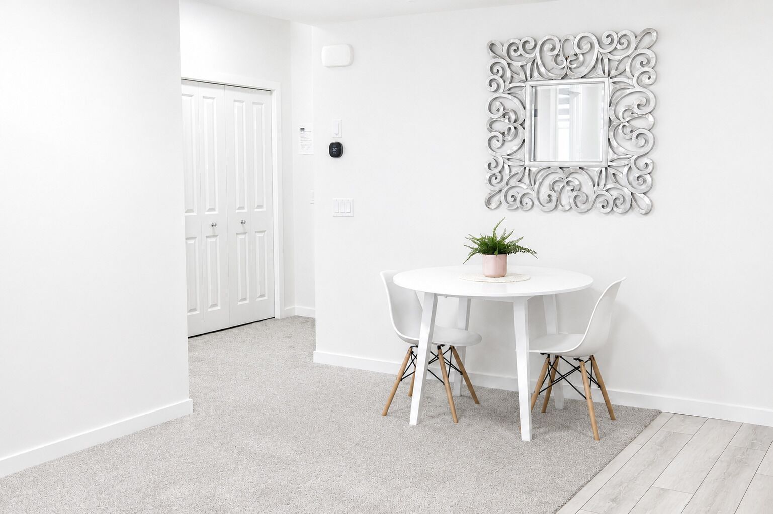 A room with a white door, a round table with two chairs, a potted plant, and a decorative mirror on the wall.