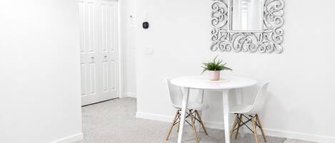 A room with a white door, a round table with two chairs, a potted plant, and a decorative mirror on the wall.