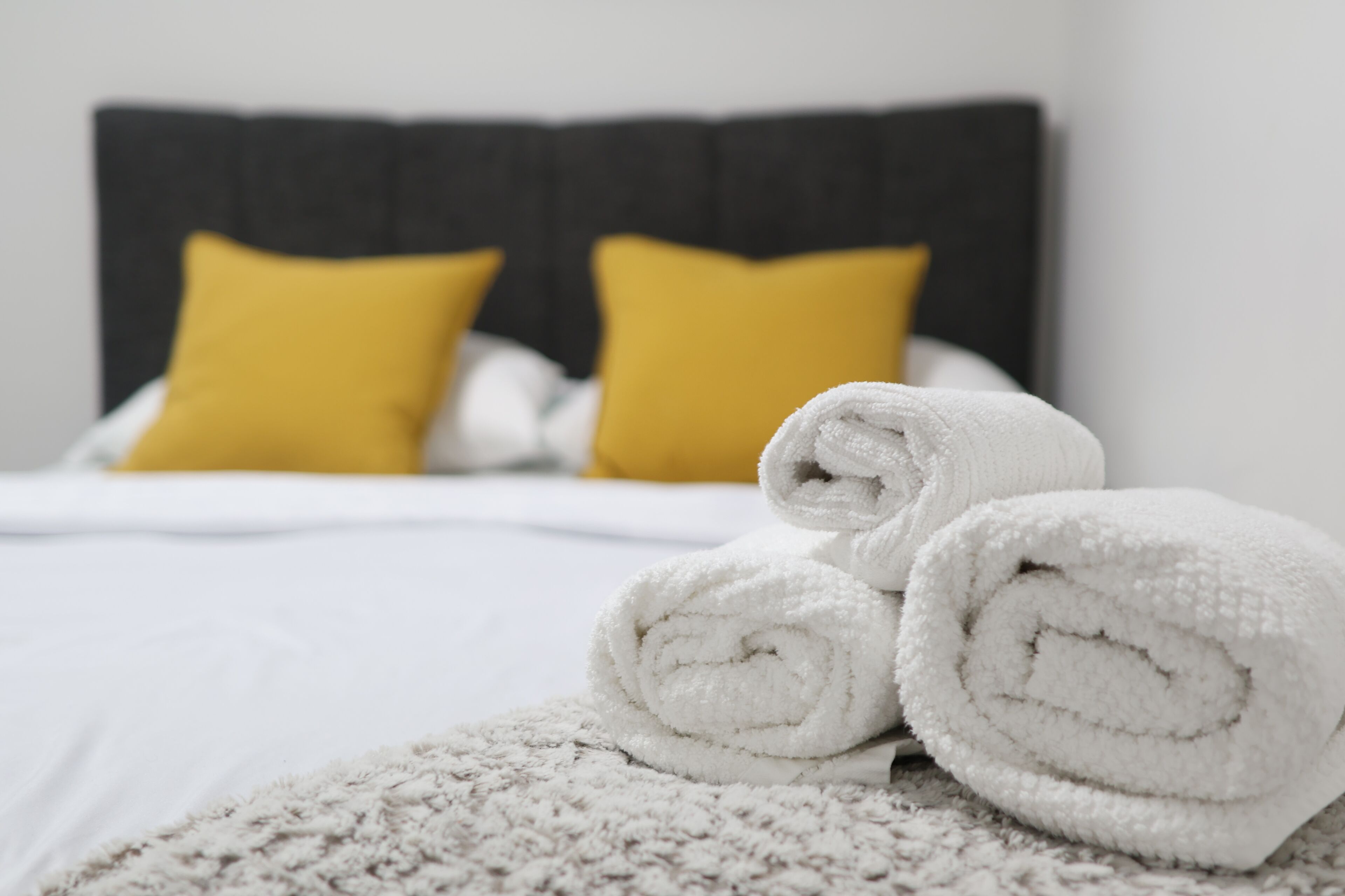A neatly made bed with white bedding and two yellow pillows, a rolled white towel on the bed, and a textured blanket.