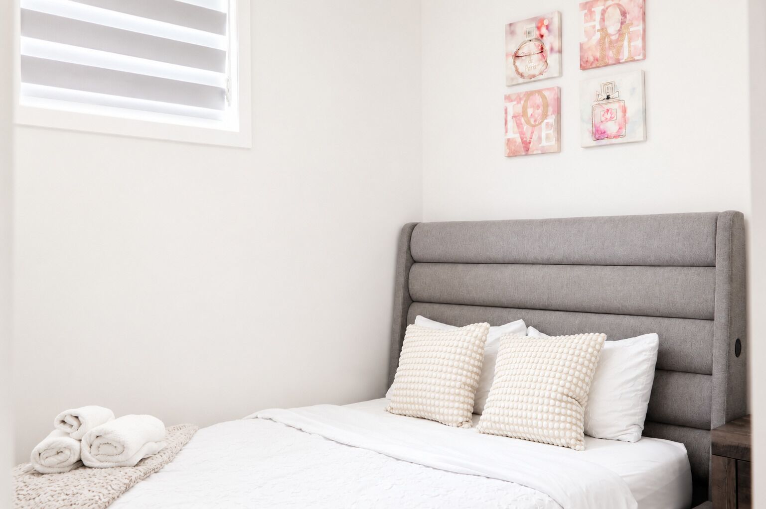 Een slaapkamer met een grijs bekleed hoofdeinde, wit beddengoed en decoratieve wanddecoratie.