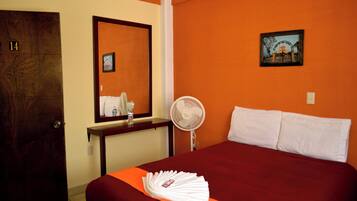 A hotel room with a bed, a bedside table, a mirror, a fan, and a door.