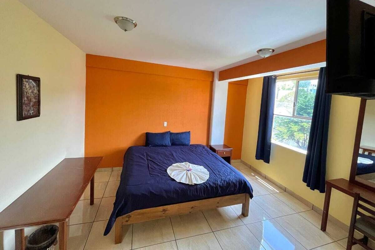 Ein Schlafzimmer mit einem Bett, einem Nachttisch aus Holz, einer Bank, einem Fernseher und einem Fenster mit Vorhängen.
