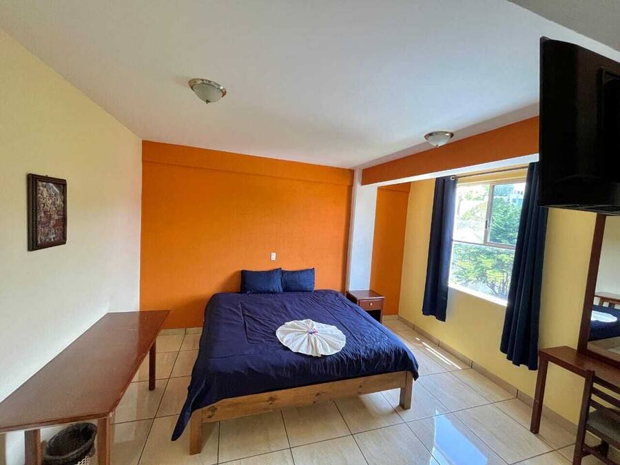 A bedroom with a bed, a wooden nightstand, a bench, a TV, and a window with curtains.