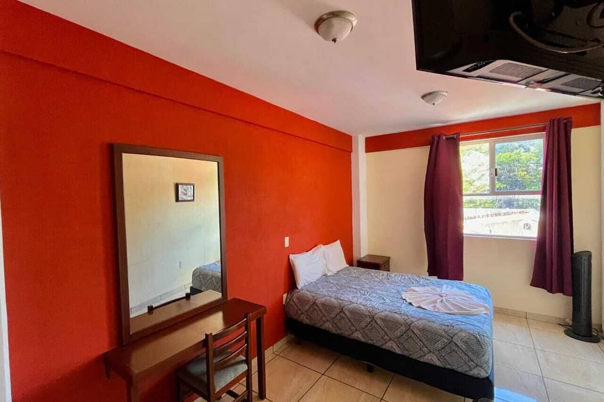 Ein Hotelzimmer mit Bett, Schreibtisch, Stuhl, einem Fenster mit Vorhängen und einem Deckenventilator.
