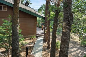A cabin with a wooden exterior, surrounded by trees and a concrete path leading to a deck. - Retro Bear Haus • Spa • Fully Fenced (Big Bear Lake)