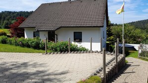 Une maison blanche avec un toit recouvert de tuiles sombres, entourée d’une allée pavée et d’une clôture en bois.