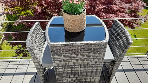 Une table d’extérieur en osier, avec un plateau en verre et un panier tressé orné de fleurs roses.