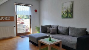 Un salon moderne avec un canapé gris, une table basse en bois et un tapis vert. Une grande fenêtre donne sur un jardin, et un panneau indiquant « HOME » est accroché au mur.