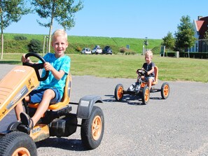 Dos niños montando tractores de juguete sobre una carretera pavimentada, con un césped y coches estacionados al fondo.
