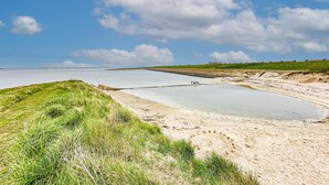 Paesaggio costiero con spiaggia sabbiosa, zone erbose e mare calmo sotto un cielo parzialmente nuvoloso.