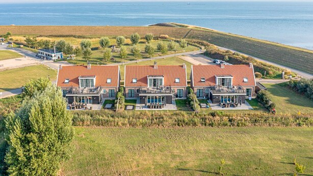 Un complesso residenziale moderno con tre case, ognuna caratterizzata da un tetto in tegole rosse e ampie finestre. Il complesso si trova vicino a un corso d'acqua, con una zona erbosa e alberi sullo sfondo.
