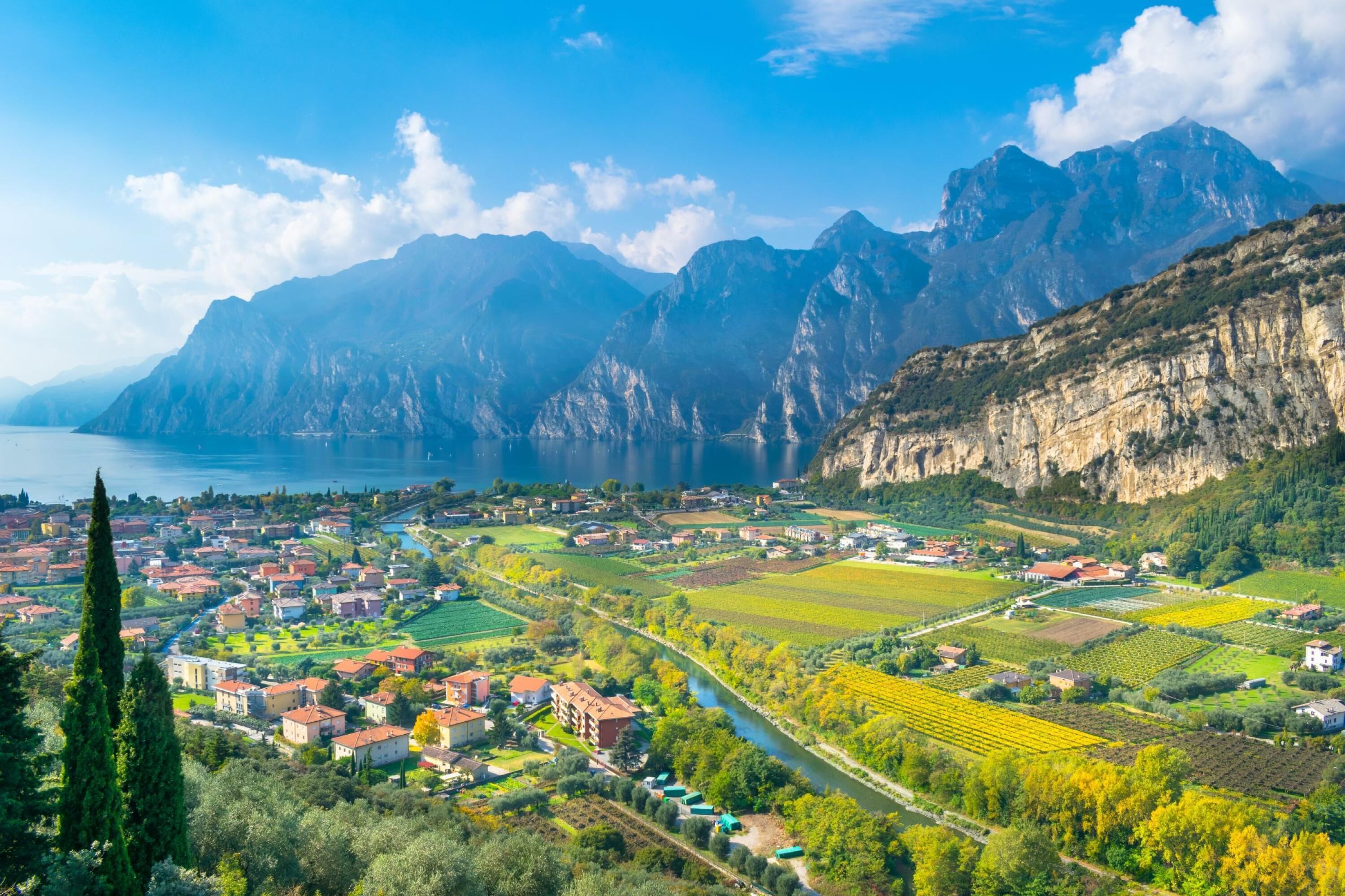 A panoramic view of a town nestled between mountains and a lake, featuring a river running through the center, surrounded by agricultural fields and residential buildings.
