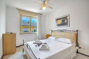 Una cama bien tendida con sábanas y toallas blancas, un ventilador de techo, una ventana con cortinas y un cuadro en la pared.