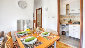 A dining area with a wooden table set for four, a clock on the wall, and a kitchen with white cabinets and a patterned backsplash.