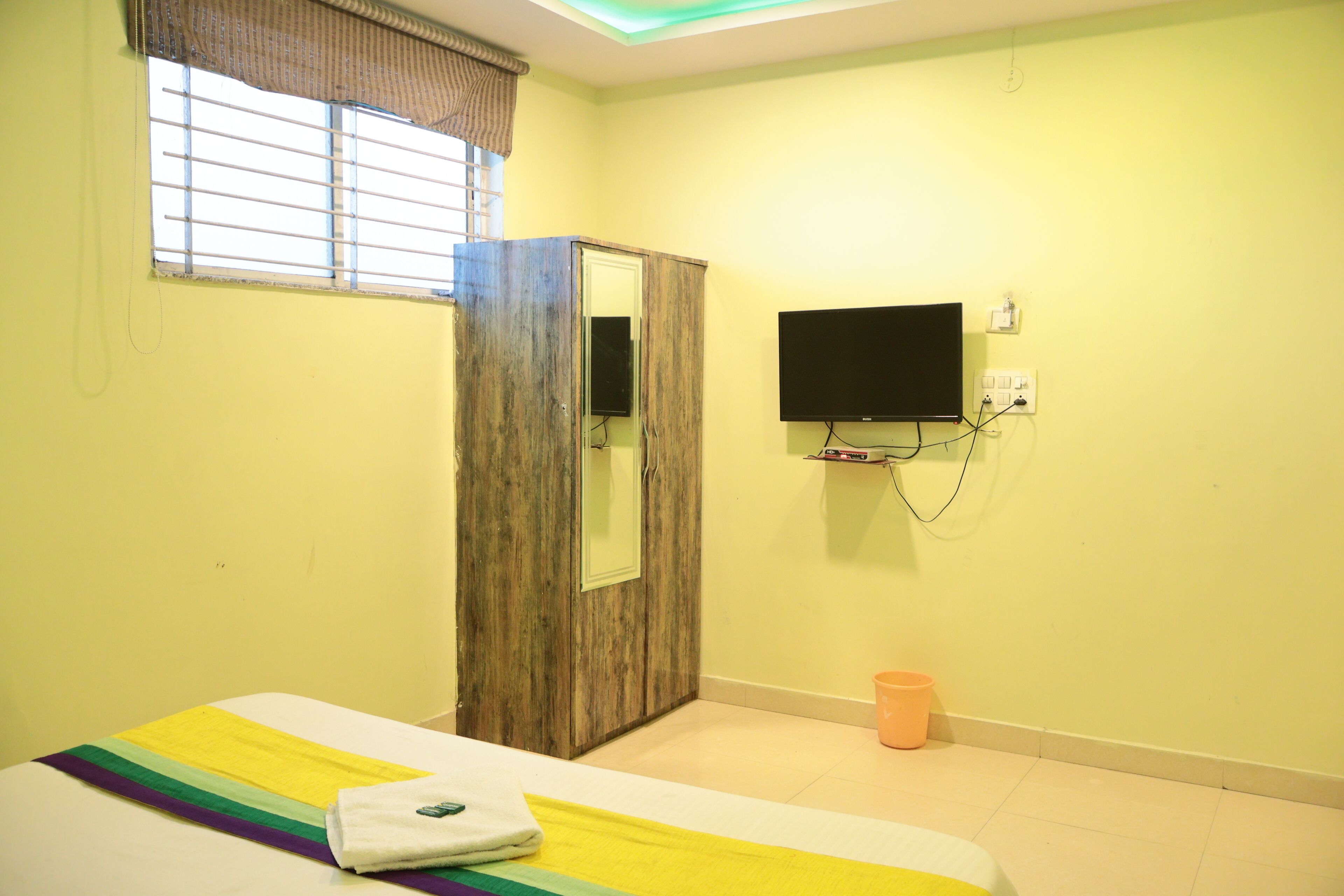 A room with a bed, a wardrobe, a television mounted on the wall, and a window with blinds.