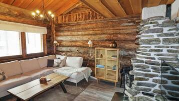 A cozy log cabin interior with a wooden ceiling, a stone fireplace, a sofa, a table, and a lamp.