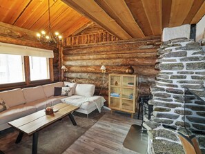 A cozy log cabin interior with a wooden ceiling, a stone fireplace, a sofa, a table, and a lamp.