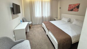 Habitación de hotel con cama, escritorio, silla, televisor y ventana con cortinas.