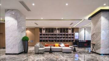 A modern lobby with a marble floor, a seating area with sofas and armchairs, a large bookshelf, and a digital screen.
