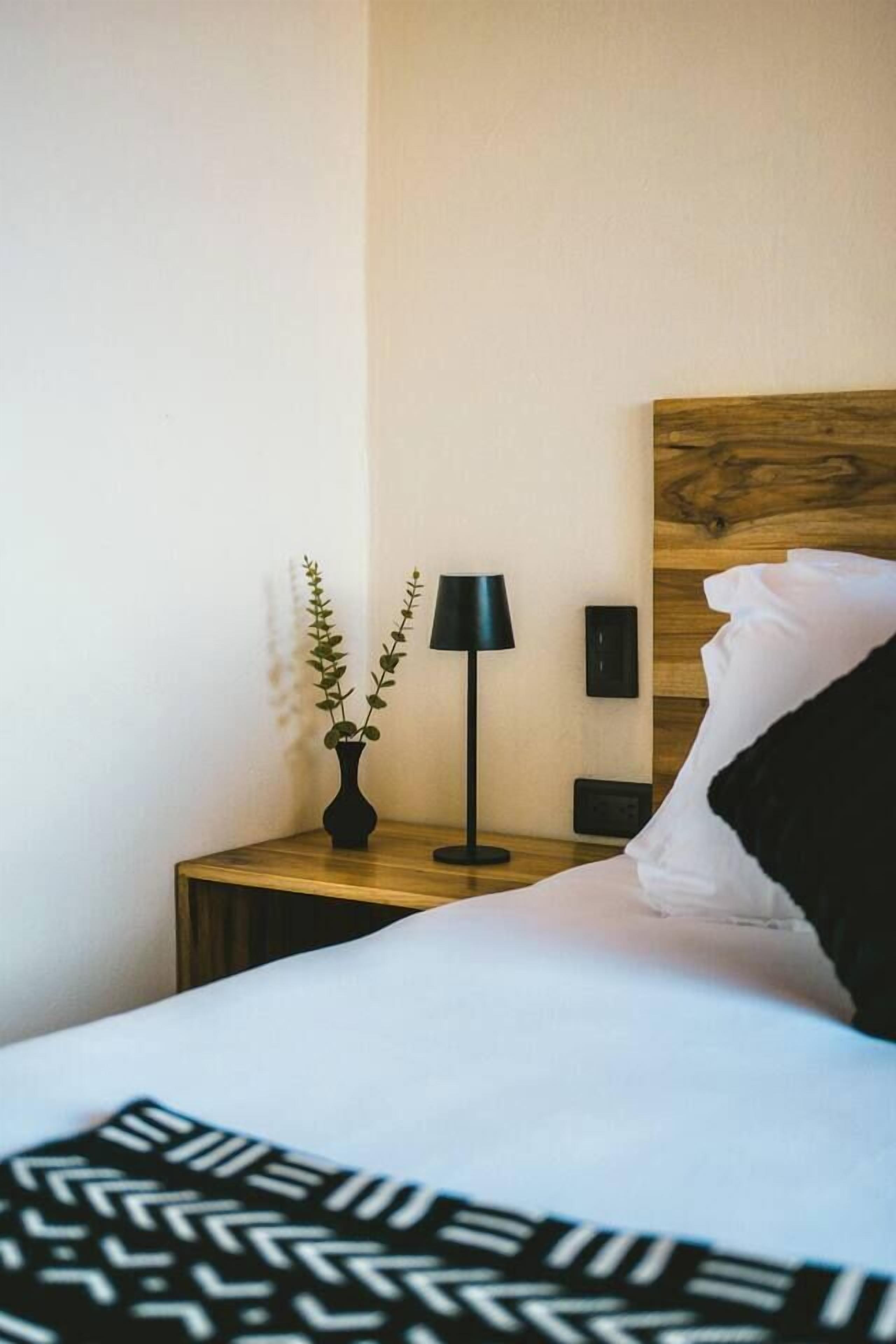 A neatly made bed with a black and white patterned bedspread, a small bedside table with a black lamp, a potted plant, and a wooden headboard.
