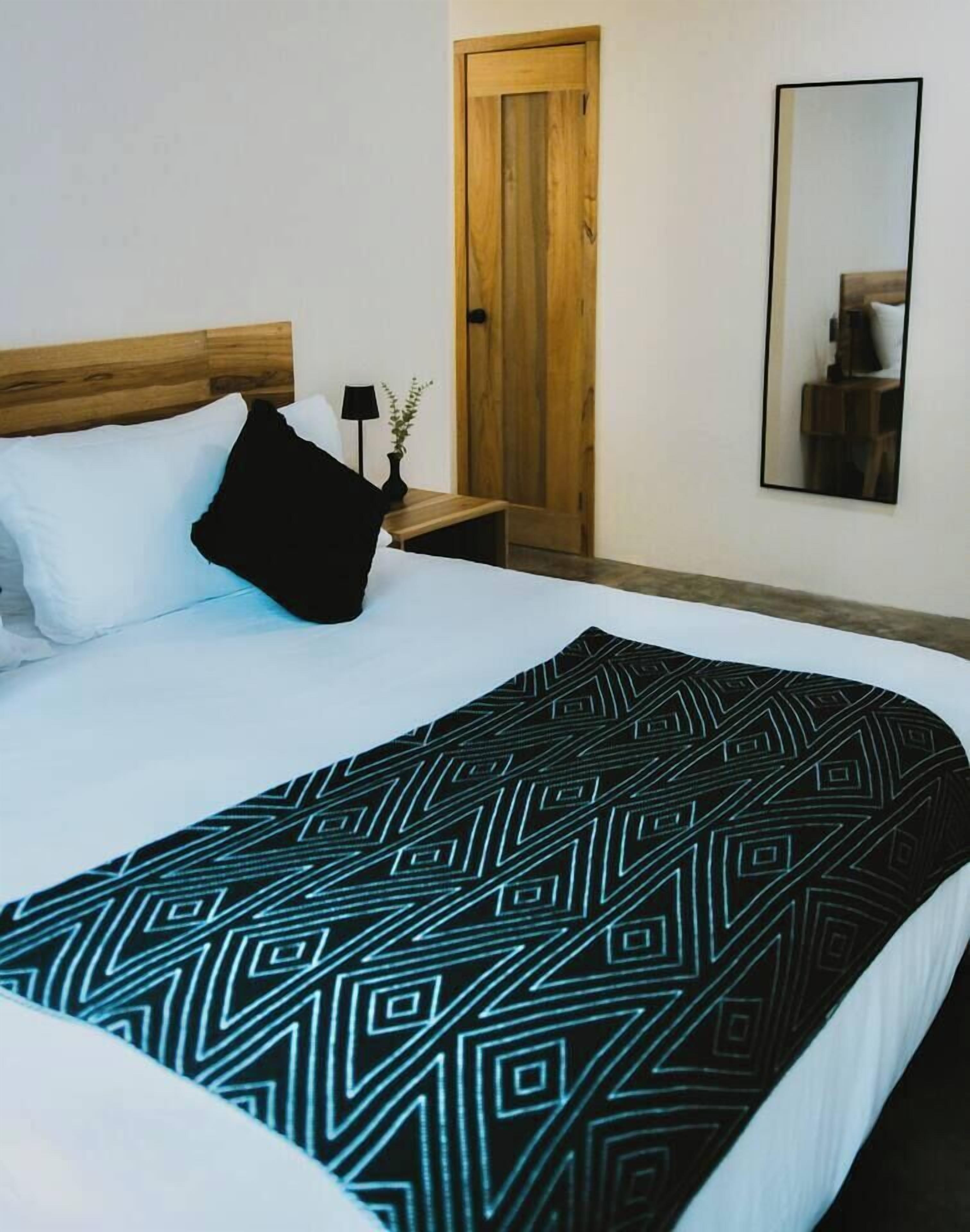 Een opgemaakt bed met een deken met een geometrisch patroon, een houten hoofdbord, een nachtkastje met een lamp en een spiegel aan de muur.