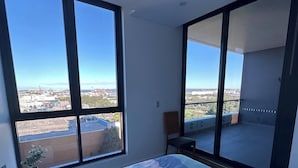 Un dormitorio con una cama grande, una silla y vistas a un estacionamiento y edificios a través de amplias ventanas.
