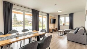 A modern living room with a dining area, large windows, a flat-screen TV, and a grey sofa. - Chalet 19 with balcony and mountain view (Inzell)