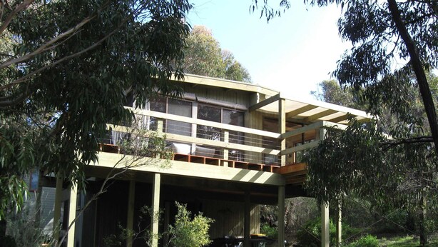 Ein zweistöckiges Holzhaus mit Balkon, umgeben von Bäumen und einem Schotterweg.