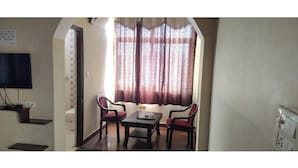 Uma área de refeições com uma mesa e duas cadeiras, uma televisão na parede e uma janela com cortinas estampadas.