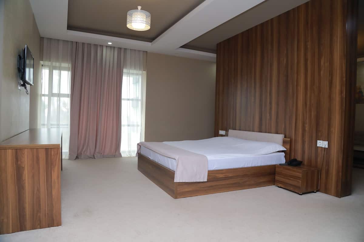 A bedroom with a wooden bed, a bedside table, a TV mounted on the wall, and a large window with curtains.