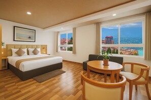 Habitación de hotel con cama, sofá, mesa de comedor y ventanas amplias con vista a la ciudad.