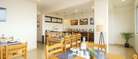 Un coin repas avec des tables et des chaises en bois, une cuisine à l’arrière-plan et une plante en pot près de la fenêtre.