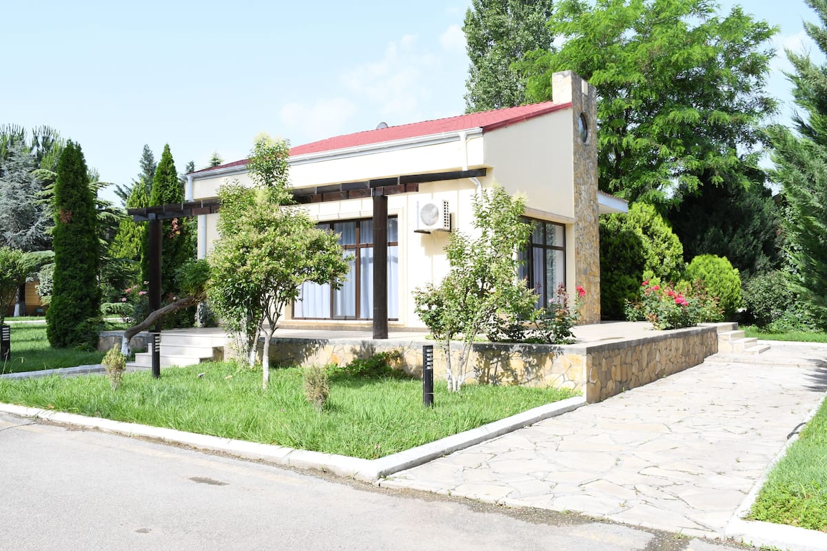 Une maison moderne à deux étages, dotée d’une façade en pierre, d’un toit rouge et d’un jardin bien entretenu où poussent diverses plantes et un petit arbre.