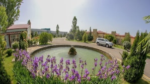 A landscaped garden with a circular pond, surrounded by purple flowers and greenery, with buildings and a parked car in the background. - Arena Hotel Gazakh (Gazakh)