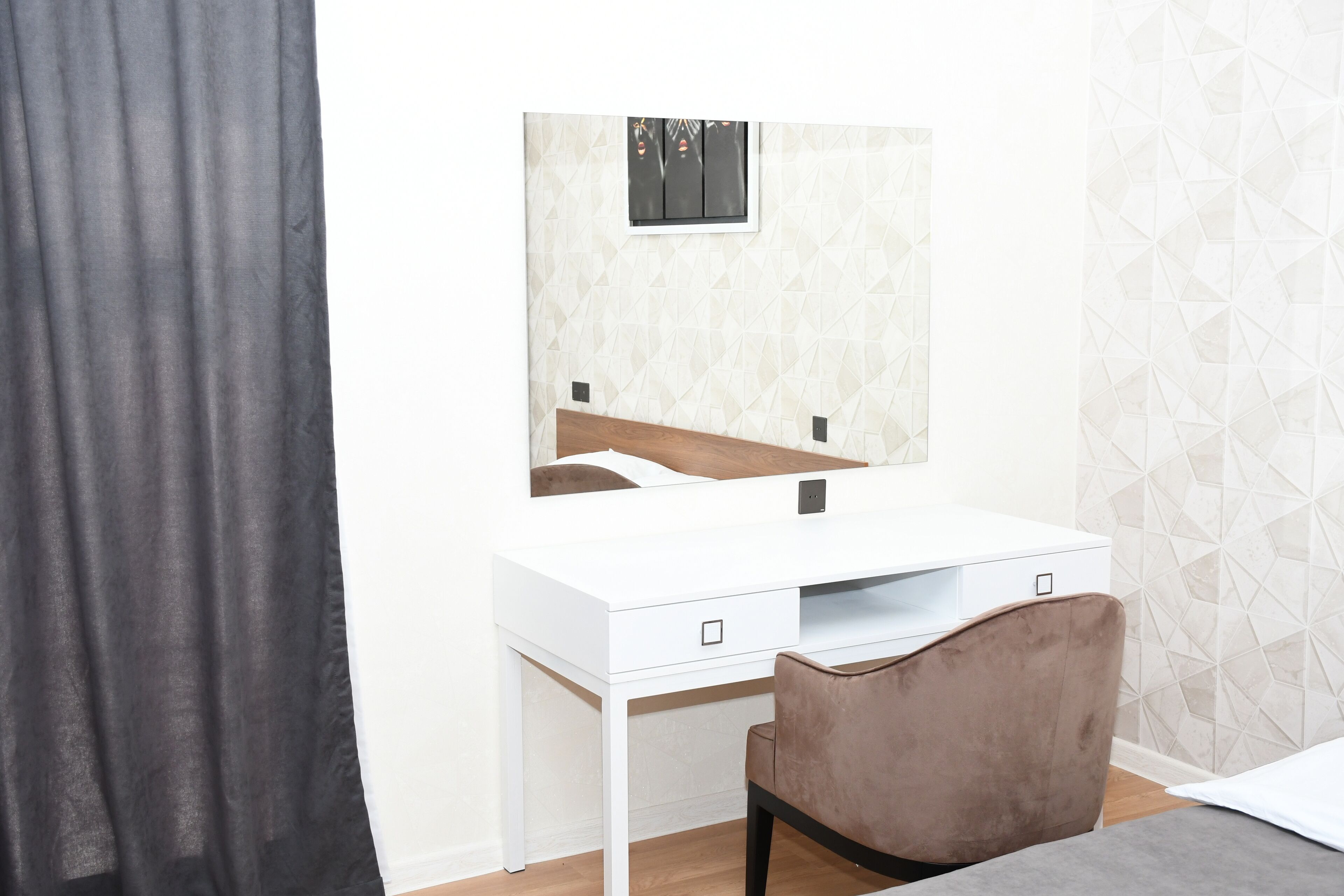 Une chambre avec un lit blanc, une chaise marron, un bureau blanc avec un miroir et un mur orné d’un motif géométrique.