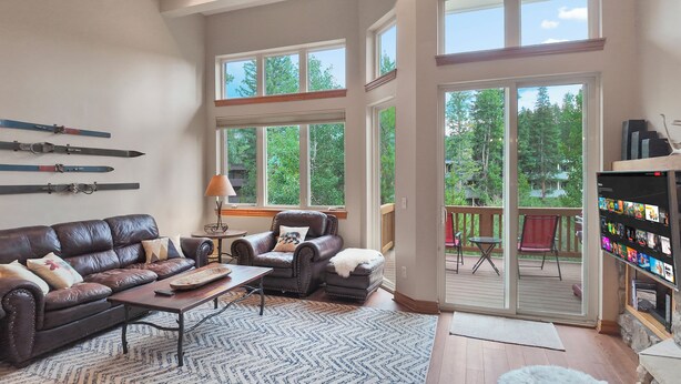 A spacious living room with a leather sofa, a coffee table, and a fireplace. There are large windows and a balcony with outdoor seating.