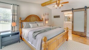 A bedroom with a wooden bed, a nightstand, a ceiling fan, and a sliding door to a bathroom. - Outdoor Kitchen + Hot Tub: Table Rock Lake House (Golden)