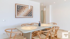 A dining area with a rectangular table, six wooden chairs, a vase with flowers, a wall-mounted artwork, and a pendant light. - Moraira Club 210 - Grupo Turis - Moraira, Costa Blanca (Moraira)