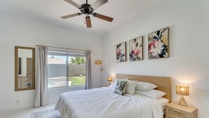 Une chambre à coucher comprenant un lit, une table de chevet, un ventilateur de plafond, une fenêtre avec des rideaux et des œuvres d’art florales accrochées au mur.