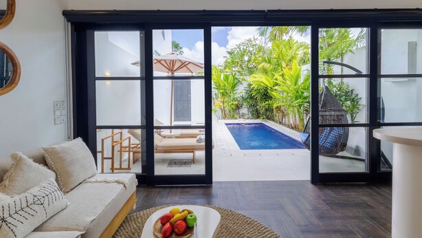 A modern living room with a sofa, a coffee table with fruit, and a view of a swimming pool and outdoor seating area through large glass doors. - Villa Gael by Pertama Stay (Seminyak)