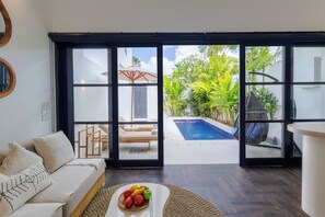 Un salon moderne avec un canapé, une table basse sur laquelle sont disposés des fruits, et une vue sur une piscine et un espace de détente extérieur, le tout visible à travers de grandes portes vitrées.
